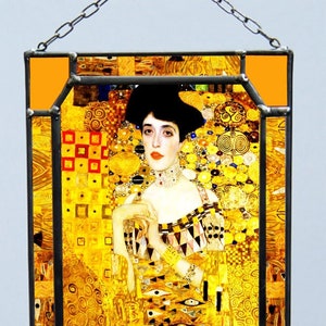 Op de afbeelding: Een gebrandschilderd raam met een portret van een vrouw op een gouden achtergrond. De vrouw draagt een zwarte hoed en een gouden ketting. De achtergrond is gemaakt van geometrische vormen en patronen.