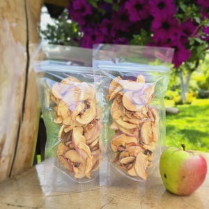 May include: Two clear plastic bags filled with dried apple slices. The bags are sitting on a wooden surface next to a green and red apple.