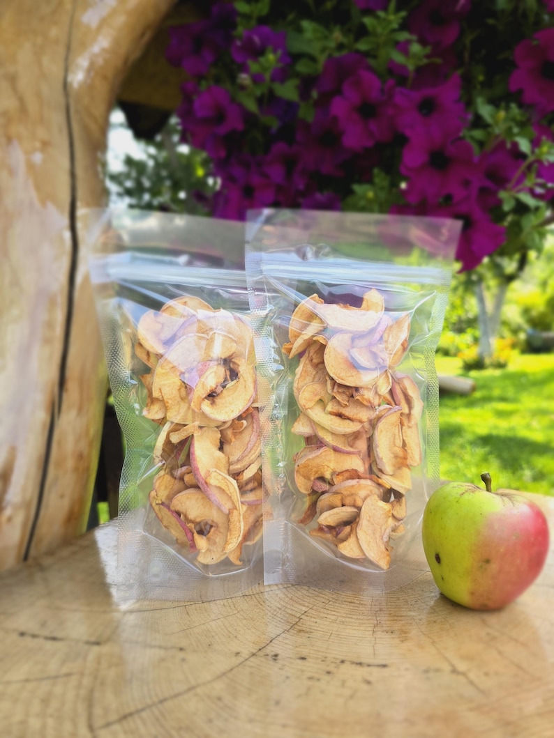May include: Two clear plastic bags filled with dried apple slices. The bags are sitting on a wooden surface next to a green and red apple.