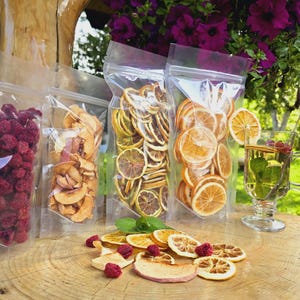 Puede incluir: Cuatro bolsas de plástico transparentes llenas de fruta seca y rodajas de cítricos. Las bolsas están sobre una superficie de madera con un vaso de té y algunas rodajas de fruta seca sueltas.