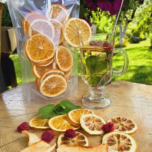 May include: A clear bag filled with dried orange slices, alongside a glass mug of tea with a lemon slice and raspberries. Fresh mint leaves and more orange slices, raspberries, and apple slices are arranged on a wooden surface.