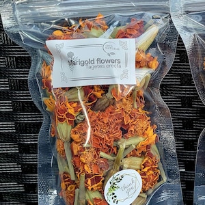 May include: A clear plastic bag filled with dried orange marigold flowers. The bag has a label that reads "100% NATURAL Marigold flowers Tagetes erecta" and a sticker that says "Thank you".