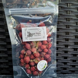 Puede incluir: Una bolsa de plástico transparente llena de escaramujos secos, etiquetada con "Rose hips Rosa canina" y "100% Natural". También hay una pequeña pegatina con las palabras "Gracias" en la bolsa.