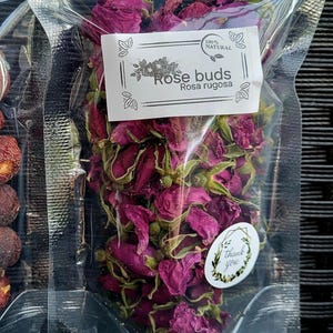 Op de afbeelding: Een doorzichtige plastic zak gevuld met gedroogde rozenknoppen. De zak heeft een label met de tekst "100% Natuurlijk Rozenknoppen Rosa rugosa".