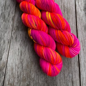 Könnte beinhalten: Zwei Knäuel buntes Garn in Pink, Orange und Gelb mit schwarzen Sprenkeln. Das Garn ist zu einer Kugel gewickelt.