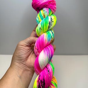 May include: A skein of hand-dyed yarn featuring vibrant neon pink, bright lime green, and electric turquoise sections. The yarn is twisted into a thick, textured bundle and includes small, scattered black speckles throughout the colourful fibres.