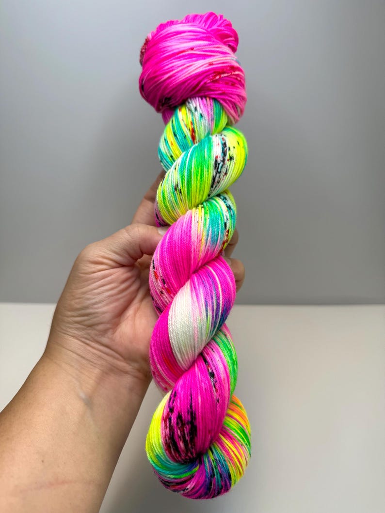 May include: A skein of hand-dyed yarn featuring vibrant neon pink, bright lime green, and electric turquoise sections. The yarn is twisted into a thick, textured bundle and includes small, scattered black speckles throughout the colourful fibres.