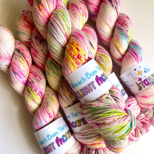 Könnte beinhalten: Drei Knäuel buntes Garn mit weißen, rosa, grünen, gelben und blauen Sprenkeln. Das Garn ist mit "Beach Bum Yarn Light Moon" beschriftet.