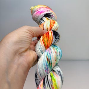 May include: A skein of white yarn featuring a vibrant multi-colored speckled pattern. The yarn displays splashes of neon pink, bright orange, electric blue, and lime green, interspersed with fine black speckles throughout the twisted fiber strands.