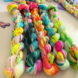 May include: Several skeins of hand-dyed yarn arranged in rows, featuring vibrant neon colors including hot pink, electric blue, lime green, and bright orange, with speckled patterns on a white base, accompanied by a small purple roller skate charm.