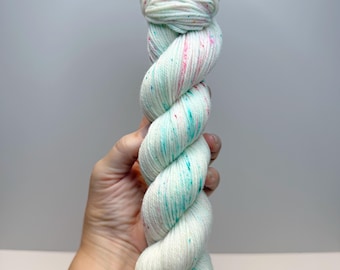 May include: A twisted skein of off-white yarn featuring a speckled pattern of small, vibrant dots in shades of turquoise, pink, and orange. The yarn has a soft, textured appearance with a consistent ply throughout the entire length of the skein.