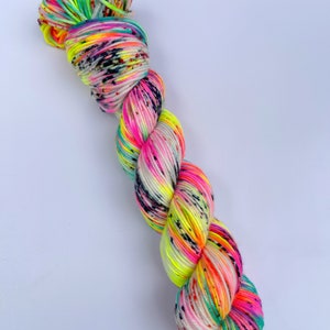 May include: A twisted skein of yarn featuring a vibrant colour palette of neon pink, bright yellow, and turquoise blue. The yarn is speckled with small black dots throughout, creating a high-contrast, multi-coloured appearance across the entire length.