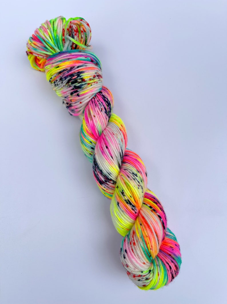 May include: A twisted skein of yarn featuring a vibrant colour palette of neon pink, bright yellow, and turquoise blue. The yarn is speckled with small black dots throughout, creating a high-contrast, multi-coloured appearance across the entire length.