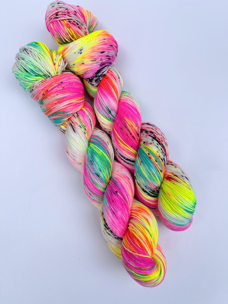 May include: Two skeins of hand-dyed yarn featuring a vibrant colour palette of neon pink, bright yellow, and turquoise green. The yarn is speckled with small black dots throughout, creating a high-contrast, multi-coloured appearance across the fibres.
