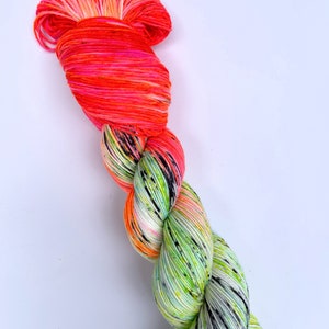 Puede incluir: Un ovillo de hilo con un degradado de color rosa brillante y naranja en la parte superior y un degradado de color verde y blanco moteado en la parte inferior.
