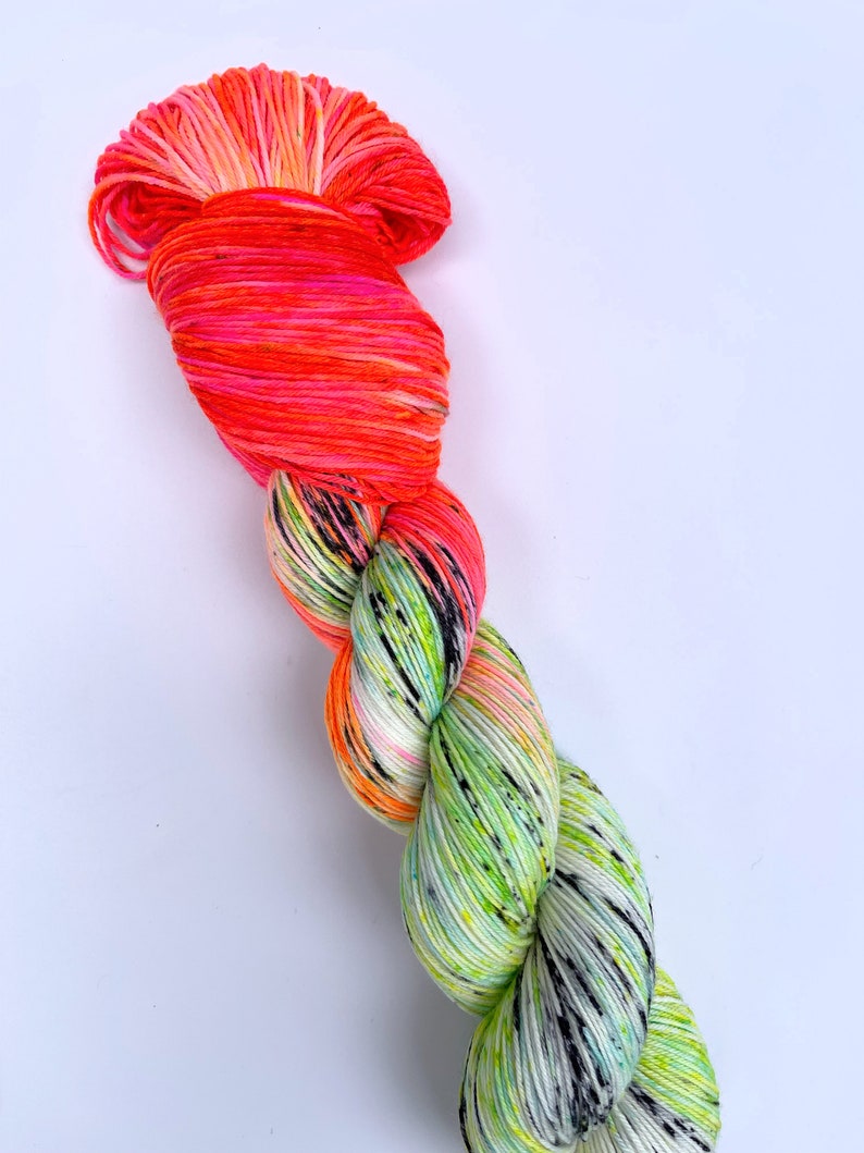 Puede incluir: Un ovillo de hilo con un degradado de color rosa brillante y naranja en la parte superior y un degradado de color verde y blanco moteado en la parte inferior.