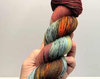 May include: A skein of hand-dyed yarn featuring a variegated colour palette of deep burgundy, burnt orange, forest green, and muted teal. The twisted fibre displays a complex blend of earthy tones with subtle speckles throughout the strands.