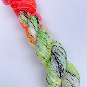 以下が含まれることがあります： 鮮やかな緑、白、黒の斑点模様に、ピンクとオレンジのグラデーションが施された手染めの糸の巻き糸です。