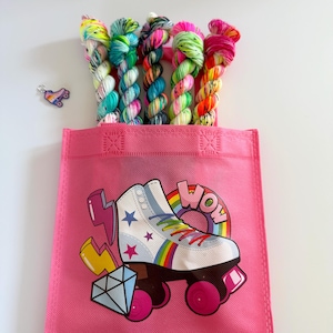 Könnte beinhalten: Eine pinkfarbene Tragetasche mit einem Rollschuh-Motiv, Regenbogen-Schnürsenkeln und dem Wort "WOW". Die Tasche enthält bunte Garnknäuel. Ein Rollschuh-Anhänger ist ebenfalls zu sehen. Die Garnknäuel sind in verschiedenen Farben.