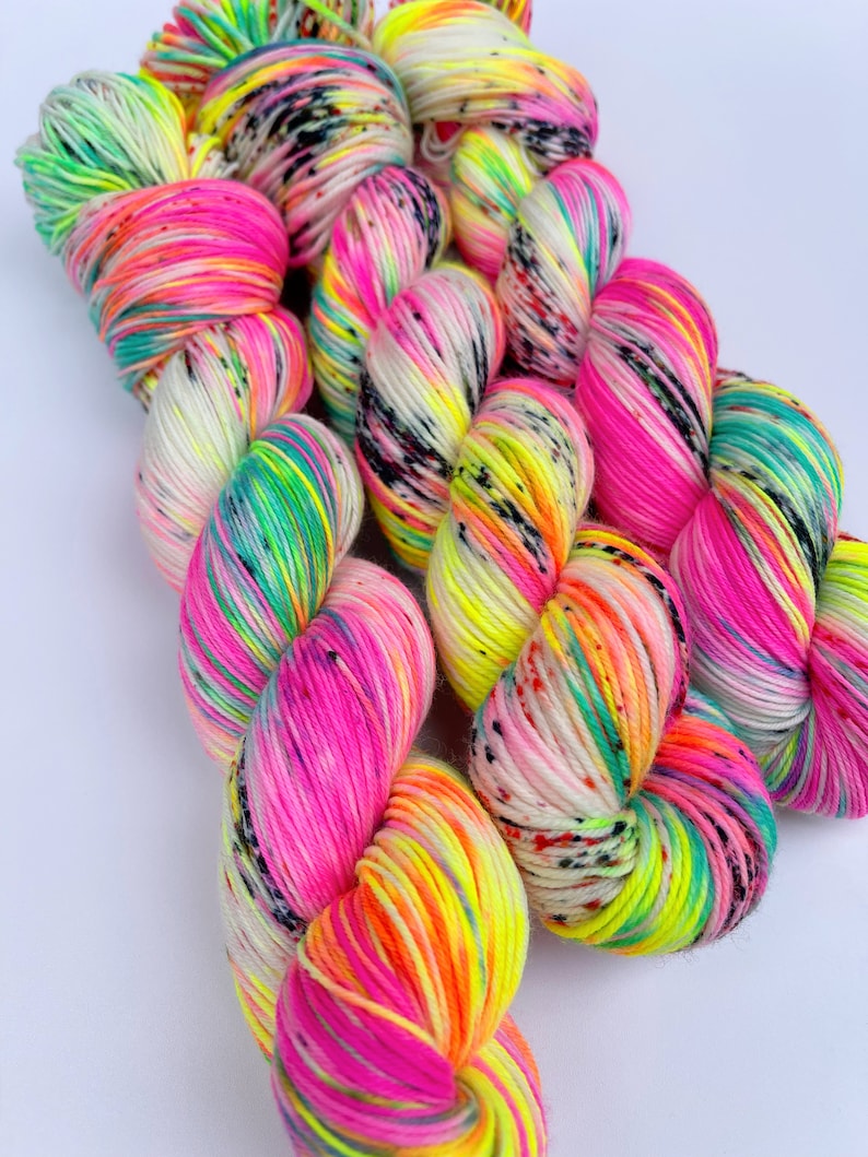May include: Three skeins of hand-dyed yarn featuring a vibrant colour palette of neon pink, bright yellow, electric green, and orange. The yarn is speckled with small black dots throughout, creating a high-contrast, multi-coloured appearance.