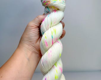 May include: A twisted skein of off-white yarn featuring vibrant, multi-coloured speckles in neon pink, bright yellow, and electric blue. The yarn has a smooth, plied texture and is presented in a long, cylindrical shape with a consistent twist.