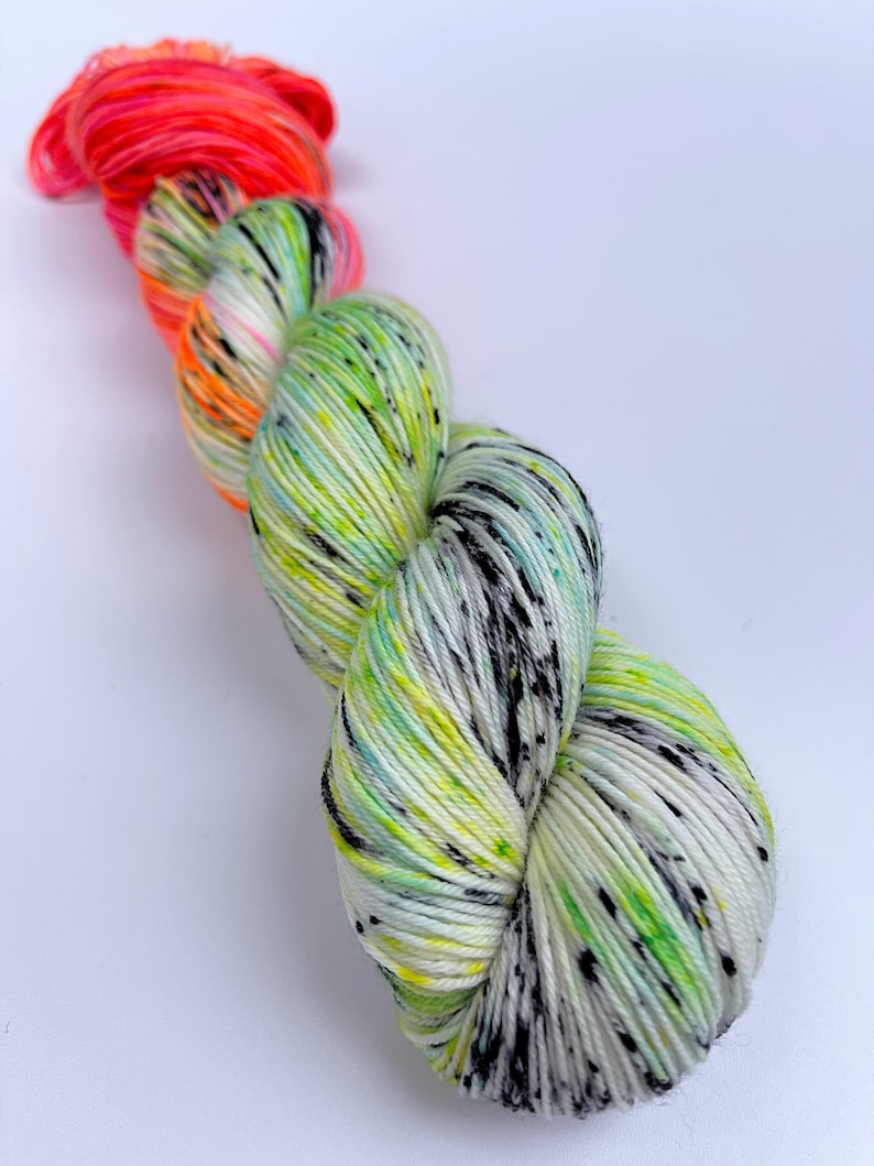 Puede incluir: Un ovillo de hilo con una base blanca y salpicaduras de verde, amarillo y negro. El hilo est&aacute; rematado con una secci&oacute;n de color rosa y naranja brillante.