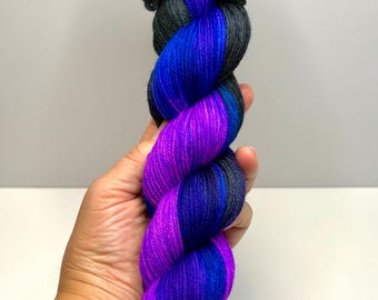 May include: A twisted skein of yarn featuring a vibrant colour gradient. The yarn transitions from a deep charcoal black at the top into alternating sections of bright royal blue and vivid magenta purple throughout the rest of the twisted fibre.