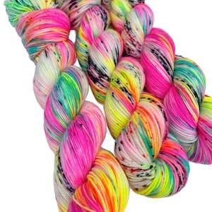 May include: Three skeins of hand-dyed yarn featuring a vibrant neon colour palette. The yarn displays a variegated pattern with bright pink, electric yellow, lime green, and turquoise hues, accented by small, scattered black speckles throughout.