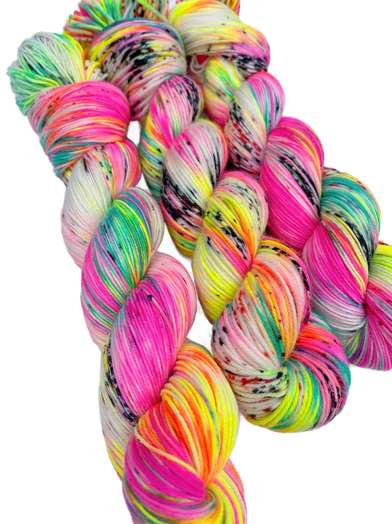May include: Three skeins of hand-dyed yarn featuring a vibrant neon colour palette. The yarn displays a variegated pattern with bright pink, electric yellow, lime green, and turquoise hues, accented by small, scattered black speckles throughout.