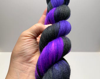 May include: A skein of yarn featuring a variegated colour pattern of deep black and vibrant purple. The fibres are twisted into a long, cylindrical shape, showcasing a consistent texture throughout the length of the yarn with alternating dark and bright sections.