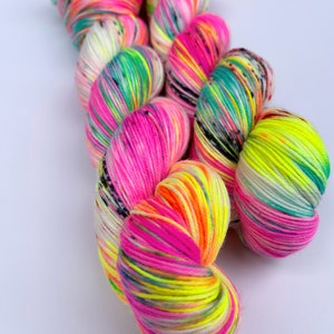 May include: A skein of hand-dyed yarn featuring a vibrant colour palette of neon pink, bright yellow, turquoise, and white. The twisted fibre displays variegated sections with small black speckles distributed throughout the multi-coloured strands.