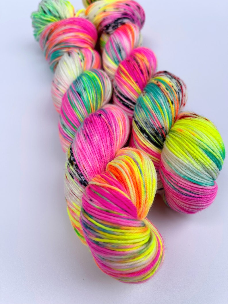 May include: A skein of hand-dyed yarn featuring a vibrant colour palette of neon pink, bright yellow, turquoise, and white. The twisted fibre displays variegated sections with small black speckles distributed throughout the multi-coloured strands.