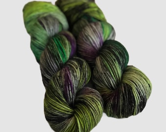 May include: Two skeins of hand-dyed yarn featuring a variegated color palette of lime green, forest green, deep purple, and charcoal gray. The twisted fiber strands display a multi-tonal appearance with blended dark and light hues throughout.
