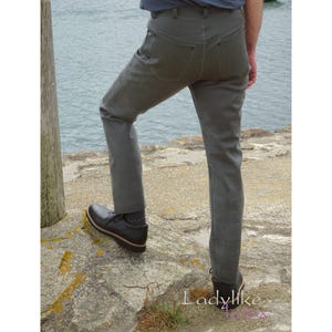 Könnte beinhalten: Graue Hose mit klassischem Fünf-Taschen-Design. Die Hose hat eine einfarbige graue Farbe und scheint aus einem strapazierfähigen Material zu bestehen. Die Person trägt schwarze Schuhe mit brauner Sohle. Der Hintergrund zeigt ein Gewässer.
