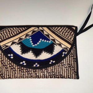 May include: A rectangular clutch with a woven straw design. The flap features a geometric pattern in black, tan, blue, and white. A black strap is attached for carrying. The bag is a stylish accessory.