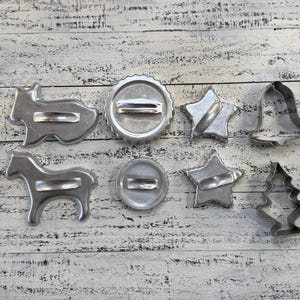 May include: A collection of vintage metal cookie cutters in various shapes, including a gingerbread man, star, bell, and horse. The cutters are arranged on a white-washed wooden surface.
