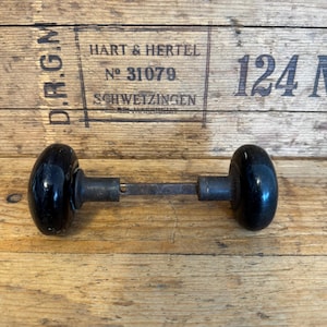 May include: Antique black door knob with a metal rod. The door knob has a rounded shape and is made of a glossy black material. The background is a wooden surface with text that reads "HART & HERTEL" and "SCHWEIZINGEN".