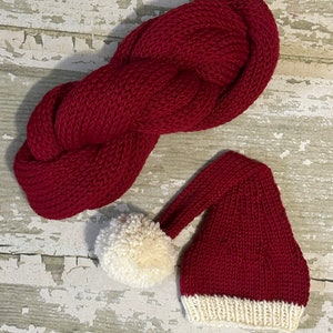 May include: A red knitted Santa hat with a white pom-pom and white trim. The hat is lying on a white wooden surface.