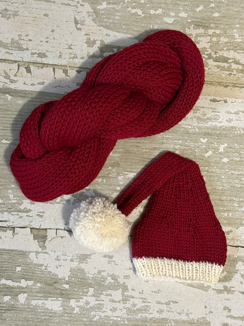 May include: A red knitted Santa hat with a white pom-pom and white trim. The hat is lying on a white wooden surface.