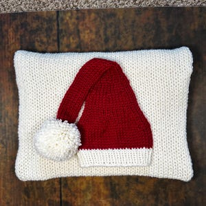 May include: A white knitted pillow with a red knitted Santa hat with a white pom-pom on top.