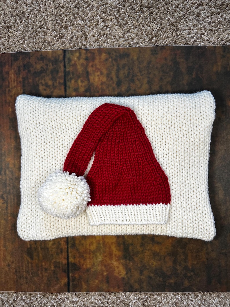 May include: A white knitted pillow with a red knitted Santa hat with a white pom-pom on top.