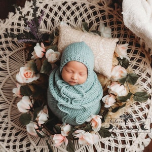May include: A newborn baby is swaddled in a light blue knitted blanket and hat, sleeping peacefully in a basket surrounded by pink roses and greenery. The basket is on a white crocheted mat.