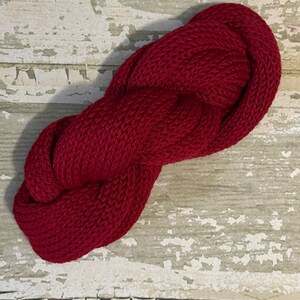 May include: A skein of deep red yarn, twisted into a figure eight shape, against a whitewashed wood background.