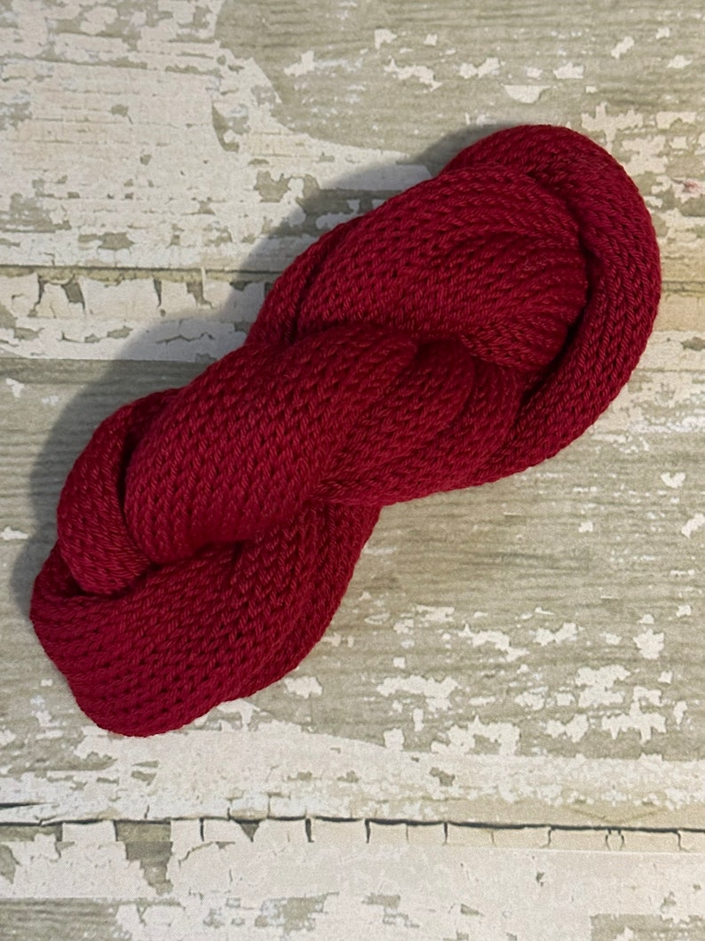 May include: A skein of deep red yarn, twisted into a figure eight shape, against a whitewashed wood background.