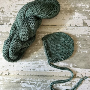May include: A green knitted baby hat and a matching knitted wrap. The hat has a long tie that can be tied under the chin.