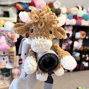 May include: A crocheted plush toy shaped like a highland cow with light brown fur, a white muzzle, white horns, and white hooves. The plush cow is positioned over the lens of a black and silver camera, featuring black plastic eyes and curly yarn.