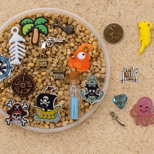 Puede incluir: Una colección de amuletos y baratijas con temática pirata, que incluyen un barco, una calavera, un ancla y una brújula. Otros amuletos incluyen una palmera, un esqueleto de pez y una pequeña botella con líquido azul.
