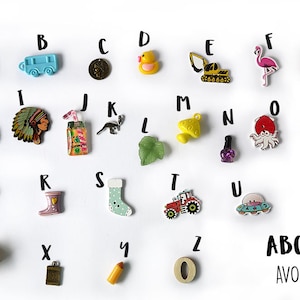 May include: A set of 26 colorful alphabet buttons with various designs, including an avocado, a bus, a coin, a rubber duck, a construction vehicle, a flamingo, a gummy bear, a hedgehog, a Native American headdress, a juice box, a kangaroo, a leaf, a mushroom, nail polish, an octopus, popcorn, a hair clip, rain boots, a sock, a tractor, a UFO, a volleyball, a waffle, a key, a pencil, and a wooden number zero. The text "ABC set AVOCADO" is printed below the buttons.