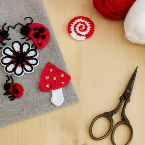 May include: Handmade crochet appliques in the image, including ladybirds, a mushroom, a flower, and a spiral design. The appliques are red, black, white, and burgundy. Vintage scissors and yarn are also visible.