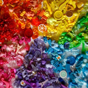 May include: A rainbow assortment of colorful plastic buttons, charms, and trinkets. The items are arranged in a rainbow pattern, starting with red and ending with blue.
